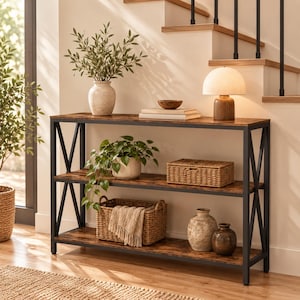 May include: A three-tier console table with a dark metal frame and brown wood-grain shelves. The top shelf holds a ceramic vase with greenery, a small bowl, books, and a lamp. The middle shelf features a potted plant and a woven basket. The bottom shelf holds a woven basket with a cloth and two ceramic vases.
