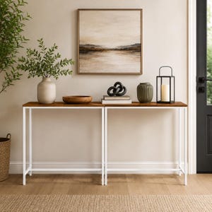 May include: A long console table with a thin white metal frame and a natural wood top. The table is decorated with a beige ceramic vase, a wooden bowl, a black sculptural knot, two stacked books, a green ribbed vase, and a black lantern.