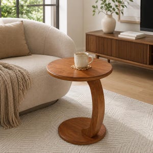 May include: A round wooden side table with a C-shaped base and a smooth, medium-brown finish. A white ceramic mug with vertical ridges sits on top of the table, resting on a small, circular, light-colored wooden coaster with a geometric pattern.