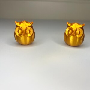 Puede incluir: Dos pequeñas figuras de búho doradas. Cada búho tiene ojos grandes y redondos y una superficie texturizada detallada. Los búhos están colocados sobre una superficie blanca, con un fondo neutro.