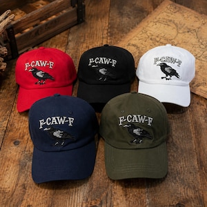 May include: Five baseball caps in red, black, white, navy blue, and olive green. Each cap features an embroidered black crow graphic and the text F-CAW-F in white or black thread. The caps have a structured crown, a curved brim, and eyelets.