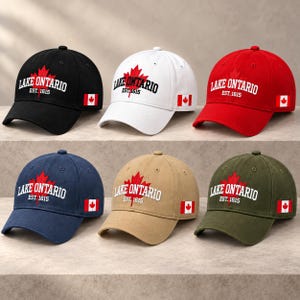 May include: Six baseball caps in black, white, red, navy blue, tan, and olive green. Each cap features an embroidered red maple leaf, the text LAKE ONTARIO EST. 1615, and a small Canadian flag patch on the side panel of the structured crown.