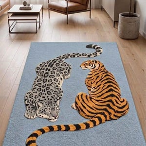 Könnte beinhalten: Ein rechteckiger Teppich in Hellblau mit einer großen, detaillierten Abbildung eines grauen Leoparden mit schwarzen Flecken, der neben einem orangefarbenen Tiger mit schwarzen Streifen auf der strukturierten blauen Oberfläche liegt.