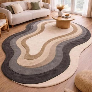 Könnte beinhalten: Ein ovaler Teppich mit einem wellenförmigen, geschichteten Muster in Creme-, Beige-, Hellgrau- und Anthrazittönen. Der Teppich hat eine weiche, strukturierte Oberfläche mit konzentrischen, organischen Kurven, die einer topografischen Karte ähneln.