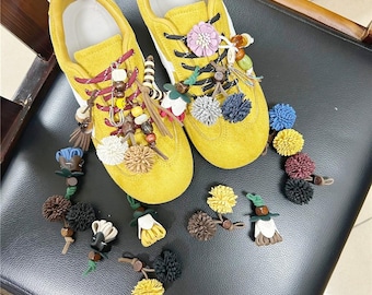 May include: A pair of bright yellow suede sneakers featuring decorative laces adorned with various colorful pom-poms, wooden beads, and small flower-shaped charms. Additional matching pom-pom and flower charms are scattered around the shoes.