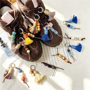 Puede incluir: Un par de zapatillas marrones decoradas con múltiples abalorios coloridos, cada uno con un anillo de metal, una pequeña borla de tela y varias cuentas de madera o plástico en tonos azul, amarillo, naranja y marrón, sobre fondo blanco.