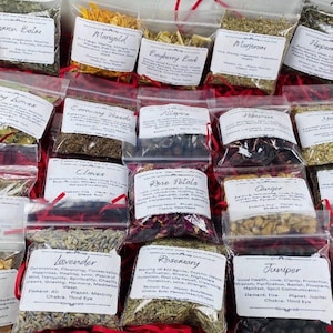 May include: A collection of small, clear plastic bags filled with various dried herbs, flowers, and roots. Each bag features a white label with printed text identifying the contents, including Lemon Balm, Marigold, Bayberry Bark, and Lavender.