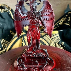 May include: A red and black resin statue of Baphomet, a goat-headed deity with wings and an upright arm. The statue has detailed features and stands on a decorative base. The background includes patterned fabric.