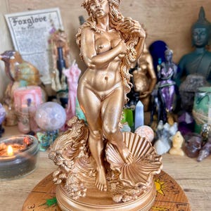 May include: A gold-colored statue of a nude woman with long flowing hair, standing on a seashell and waves. The statue is on a wooden base with painted symbols. Other decorative items are in the background.
