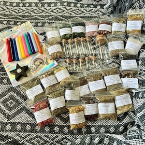 May include: A collection of dried herbs and botanicals in clear plastic pouches, small glass vials, and a larger glass jar. Includes a set of colourful taper candles, a black star-shaped candle holder, and silver-toned metal charms.