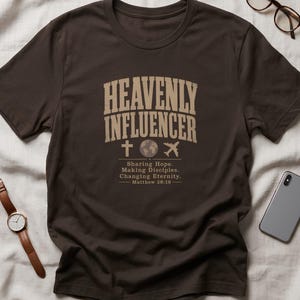 Könnte beinhalten: Ein dunkelbraunes Kurzarm-T-Shirt mit dem Text HEAVENLY INFLUENCER in einer beigen, gebogenen Schriftart. Unter dem Text befinden sich Symbole eines Kreuzes, eines Globus und eines Flugzeugs, gefolgt von Sharing Hope. Making Disciples. Changing Eternity. Matthew 28:19.