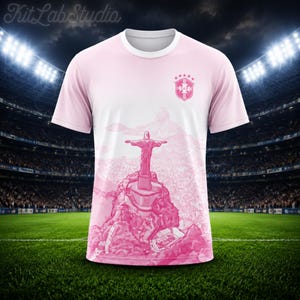 May include: A short-sleeve t-shirt in shades of pink and white featuring a graphic illustration of the Christ the Redeemer statue in Rio de Janeiro. The upper chest displays a crest with five stars, and the lower section shows a detailed landscape.