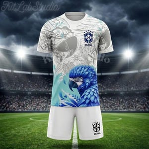 Könnte beinhalten: Ein weißes Fußballtrikot-Set mit Shorts, das mit einer blauen Ara-Grafik verziert ist. Das Trikot zeigt das blau-weiße CBF Brasil-Wappen auf der Brust, während die Shorts ein passendes CBF Brasil-Logo auf dem linken Bein aufweisen.
