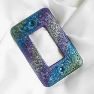 May include: A decorative light switch plate with a rectangular opening and two screw holes. The plate features a swirling, textured pattern in shades of blue, green, purple, and silver, creating an iridescent effect. The plate is set against a white background.