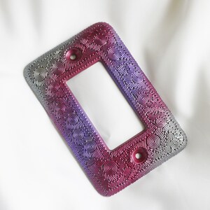 May include: A decorative light switch cover with a silver and purple swirl pattern. The cover is made of a textured material and has a rectangular shape.