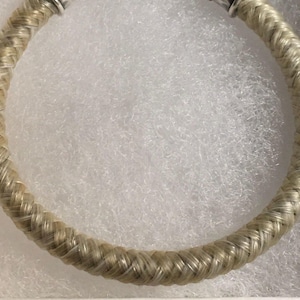May include: A circular bracelet featuring a braided, light-colored, fibrous texture. The band is constructed with a consistent woven pattern throughout its length, terminating in metallic silver-toned end caps at the top opening of the piece.