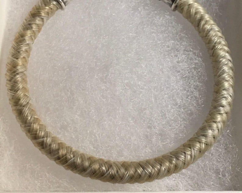 May include: A circular bracelet featuring a braided, light-colored, fibrous texture. The band is constructed with a consistent woven pattern throughout its length, terminating in metallic silver-toned end caps at the top opening of the piece.