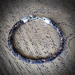 May include: A braided bracelet made of dark brown and black hair strands, featuring silver-toned metal end caps and a lobster claw clasp. The circular accessory displays a tight, uniform weave pattern throughout its length, secured by metallic hardware.