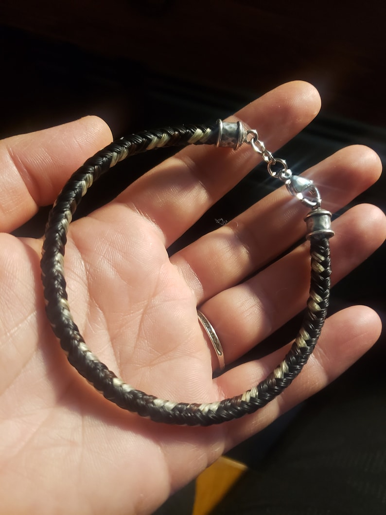 May include: A braided bracelet featuring dark brown and light blonde strands woven together in a tight pattern. The bracelet is finished with silver-toned metal end caps and a lobster claw clasp, creating a secure closure for the circular band.