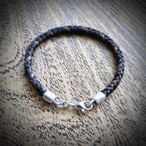 May include: A circular bracelet crafted from braided dark brown and black hair strands. The ends of the braid are secured with smooth silver-toned metal caps, and the piece is fastened with a matching silver-toned lobster claw clasp.