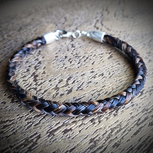 May include: A braided bracelet crafted from strands of dark brown, black, and light brown hair. The circular band features a polished silver-toned metal clasp at the top, connecting the two ends of the braided hair into a continuous loop.