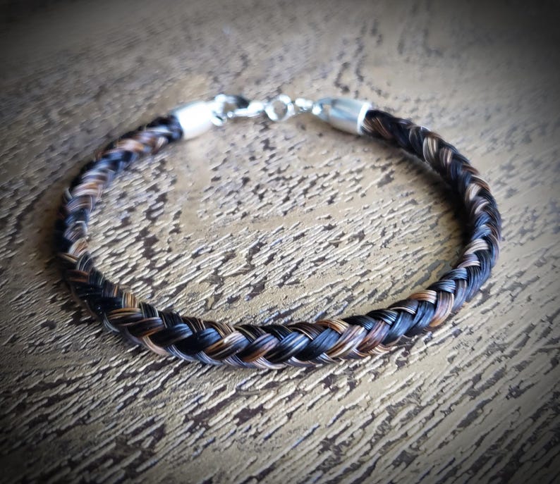 May include: A braided bracelet crafted from strands of dark brown, black, and light brown hair. The circular band features a polished silver-toned metal clasp at the top, connecting the two ends of the braided hair into a continuous loop.