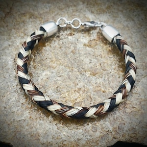 May include: A braided bracelet crafted from strands of black, white, and brown hair. The circular band features polished silver-toned metal end caps and a matching lobster claw clasp mechanism to secure the jewelry piece around the wrist.