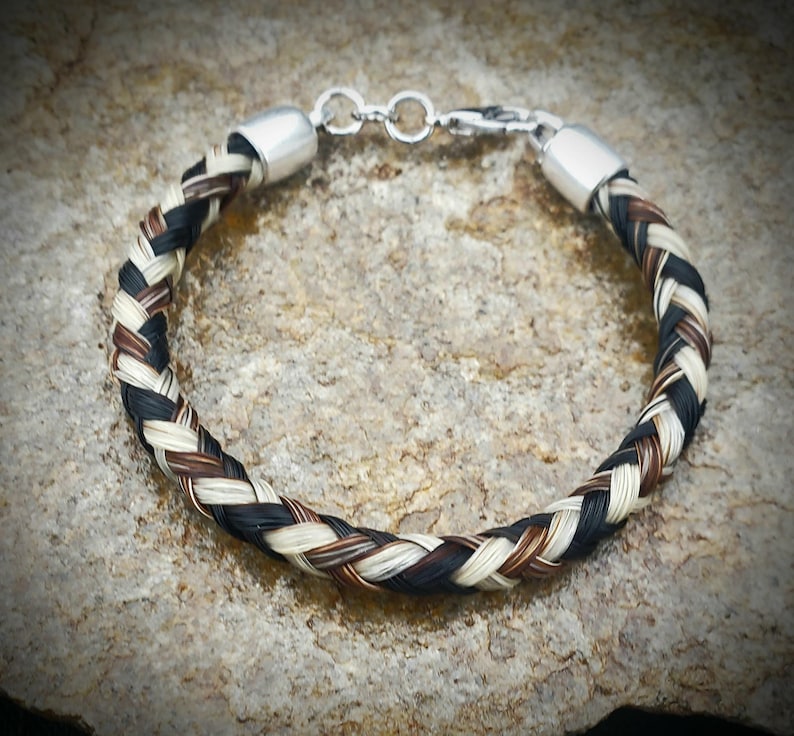 May include: A braided bracelet crafted from strands of black, white, and brown hair. The circular band features polished silver-toned metal end caps and a matching lobster claw clasp mechanism to secure the jewelry piece around the wrist.