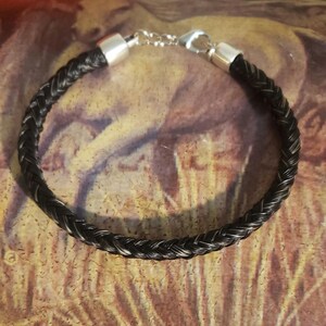 May include: A braided bracelet made of dark black and grey hair strands. The bracelet features polished silver-toned metal end caps and a silver-toned lobster claw clasp with a short extender chain, creating a circular band design.