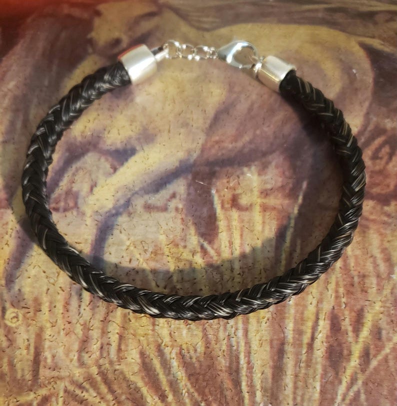 May include: A braided bracelet made of dark black and grey hair strands. The bracelet features polished silver-toned metal end caps and a silver-toned lobster claw clasp with a short extender chain, creating a circular band design.