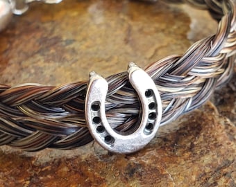 May include: A bracelet crafted from braided brown, black, and white horsehair, featuring a silver-toned metal horseshoe charm at the center. The bracelet includes silver-toned metal end caps and a lobster claw clasp with a short extender chain.