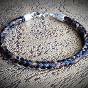 May include: A braided bracelet crafted from dark brown and black hair strands, featuring polished silver-toned metal end caps and a secure lobster claw clasp mechanism. The circular accessory displays a tight, uniform weave pattern throughout.