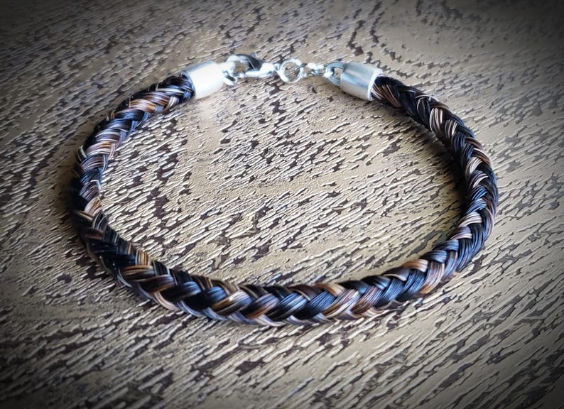 May include: A braided bracelet crafted from dark brown and black hair strands, featuring polished silver-toned metal end caps and a secure lobster claw clasp mechanism. The circular accessory displays a tight, uniform weave pattern throughout.
