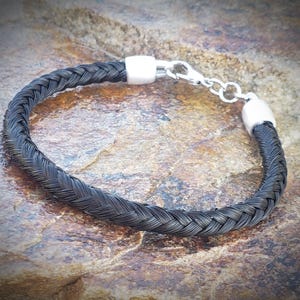 May include: A bracelet featuring a dark, braided strand of hair or fiber, secured at both ends by polished silver-toned metal caps. The piece includes a silver-toned lobster claw clasp and a short extender chain for adjustable closure.