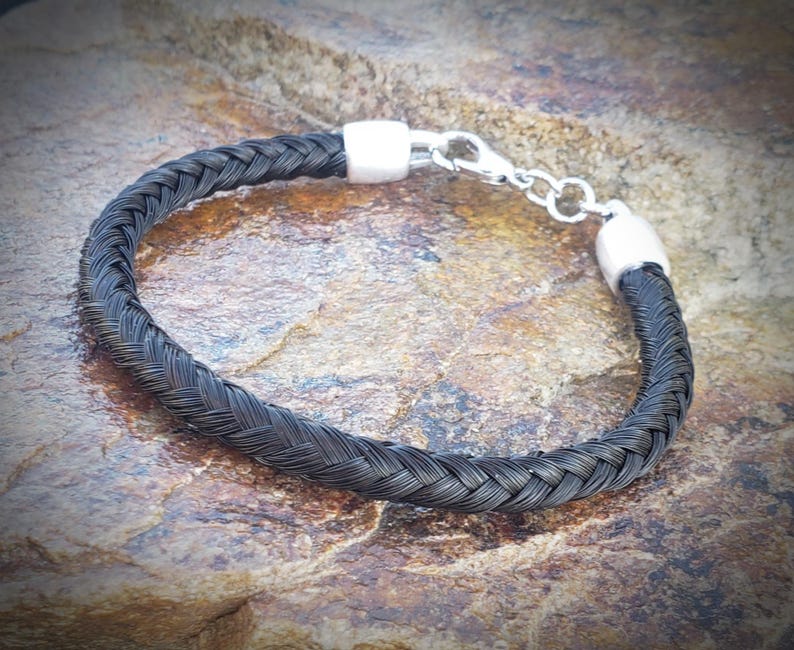 May include: A bracelet featuring a dark, braided strand of hair or fiber, secured at both ends by polished silver-toned metal caps. The piece includes a silver-toned lobster claw clasp and a short extender chain for adjustable closure.
