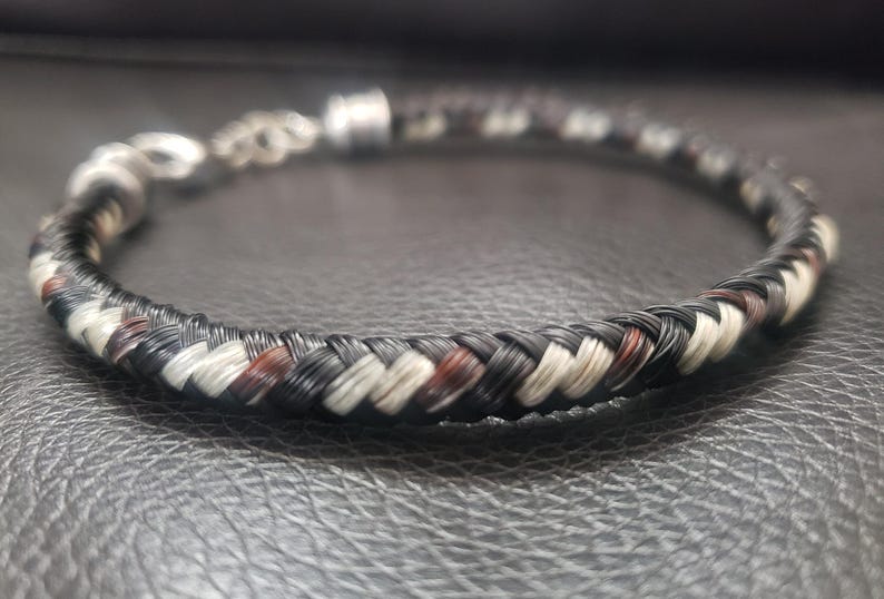 May include: A braided bracelet featuring a pattern of black, white, and brown strands. The circular band includes a silver-toned metal clasp and an adjustable chain link extension, showcasing a detailed woven texture throughout the entire piece.
