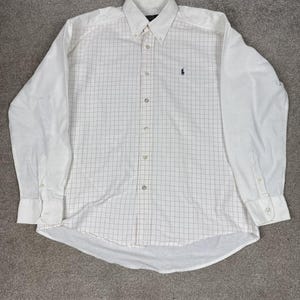 May include: White button-down shirt with a grid pattern and long sleeves. The shirt features a small, dark blue embroidered logo on the chest. The collar is button-down, and the cuffs have button closures. The shirt is laid flat on a gray surface.
