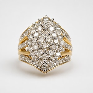 Pode incluir: Um anel tipo cluster com uma grande disposição oval pontiaguda de numerosas pedras pequenas, redondas e claras cravadas em metal branco, montado numa banda de ouro amarelo polido que se divide em três ramos incrustados de diamantes.