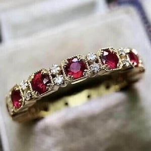 May include: A gold band ring featuring a series of alternating round-cut red rubies and small clear white diamonds. The gemstones are set in a repeating pattern along the top of the metal band, which has a polished finish and a textured edge.