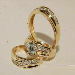 May include: A set of three gold rings featuring a polished finish. One ring displays a prominent round-cut clear gemstone held by prongs, while the other two bands are inlaid with a row of small, sparkling clear stones along the centre channel.