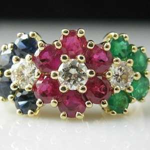 May include: A gold ring featuring a central cluster of round-cut red rubies surrounding a clear white diamond. To the left is a cluster of dark blue sapphires and a clear diamond, and to the right is a cluster of green emeralds and a clear diamond.