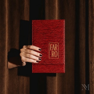 May include: A rectangular, deep red menu cover with a textured, wood-grain-like surface. The word FARO is printed in gold, capitalized letters inside a thin gold rectangular border on the front cover. The item has stitched edges.
