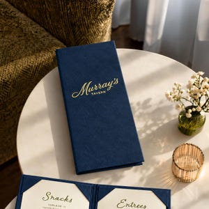 May include: A rectangular navy blue menu cover with a textured surface and the gold embossed text Murray's Tavern. Below it, an open menu displays white pages with the headings Snacks and Entrees in a gold script font, held by blue corners.