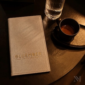 Puede incluir: Una funda de menú rectangular de tela gris claro con el texto dorado en relieve BLUEMBER NEW AMERICAN RESTAURANT, colocada junto a una taza de cerámica negra llena de café oscuro y un vaso transparente estriado sobre una superficie oscura.