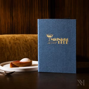 Könnte beinhalten: Ein blaues, strukturiertes Menücover mit goldener Aufschrift MidNight BLUE und einem Cocktailglas-Symbol. Daneben steht ein weißer Teller mit einem braunen, geschichteten Dessert, das mit einem Klecks weißer Creme garniert ist.