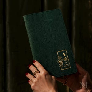 May include: A dark green menu cover with a textured finish. It has a gold rectangular emblem with floral illustrations and Chinese characters. The text "MOLI RESTAURANT" is in gold. The menu is held by a person with red nail polish.