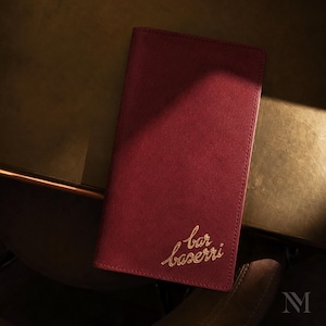 May include: A rectangular, deep red, suede-textured bill presenter or menu cover featuring the embossed gold script text "bar baserri" in the bottom right corner. The item has clean, stitched edges and a smooth, matte finish throughout.