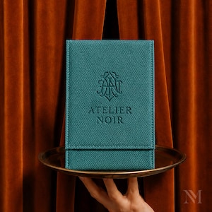 May include: A rectangular teal case with a textured fabric finish featuring the embossed logo and text ATELIER NOIR. The case has a flap closure and is positioned on a round metallic tray. The design is centered and upright with visible stitching.