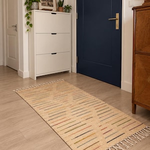 Peut inclure: Un tapis rectangulaire avec une base beige clair et un motif répétitif en losange formé par de fines lignes horizontales de différentes couleurs, dont le rouge, le bleu, le vert et le jaune. Le tapis présente des franges blanches nouées.