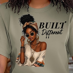 May include: An olive green t-shirt featuring a graphic of a person with a high bun, wearing sunglasses, a headwrap, and layered necklaces. The text "BUILT Different" is printed in black next to the illustration, which includes a cream-colored top.